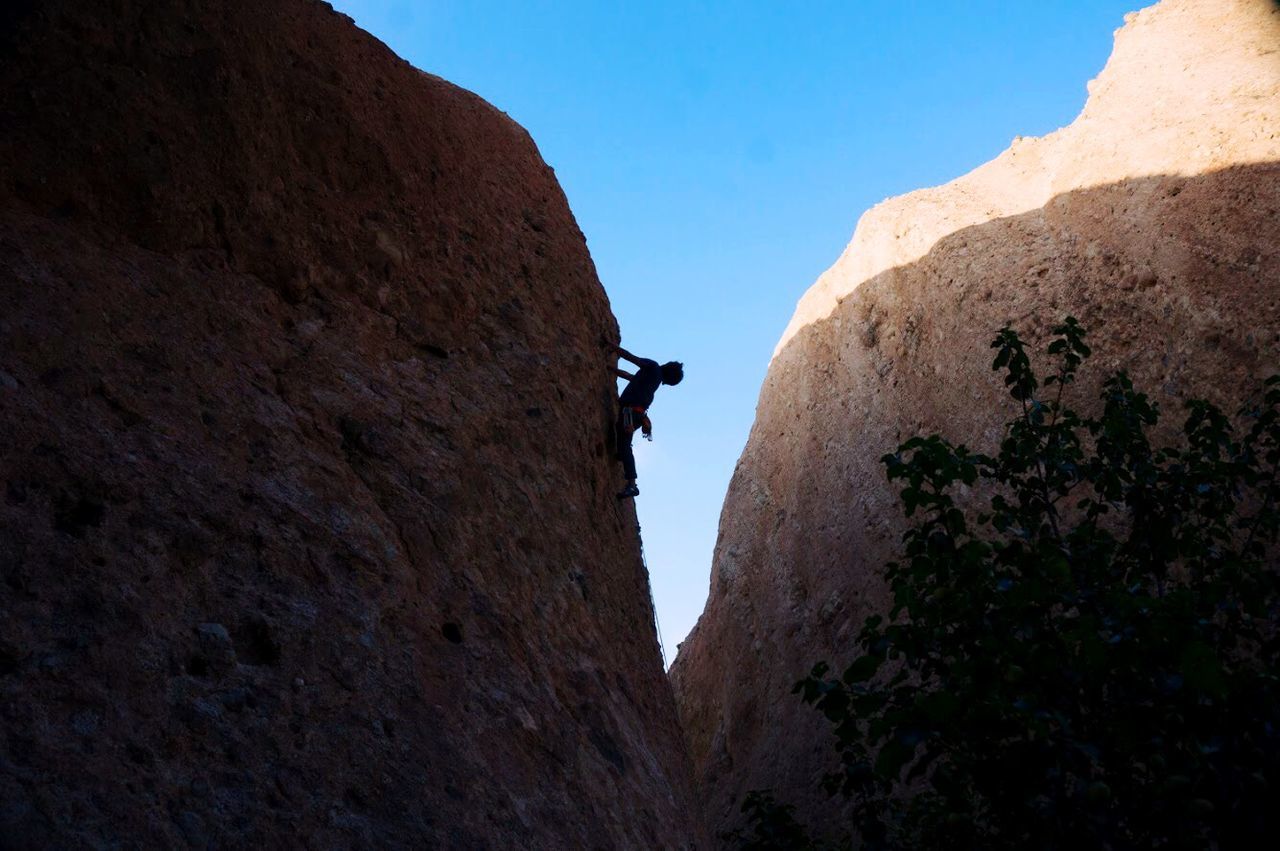 Low angle view of silhouette man climbing rocks against clear blue sky