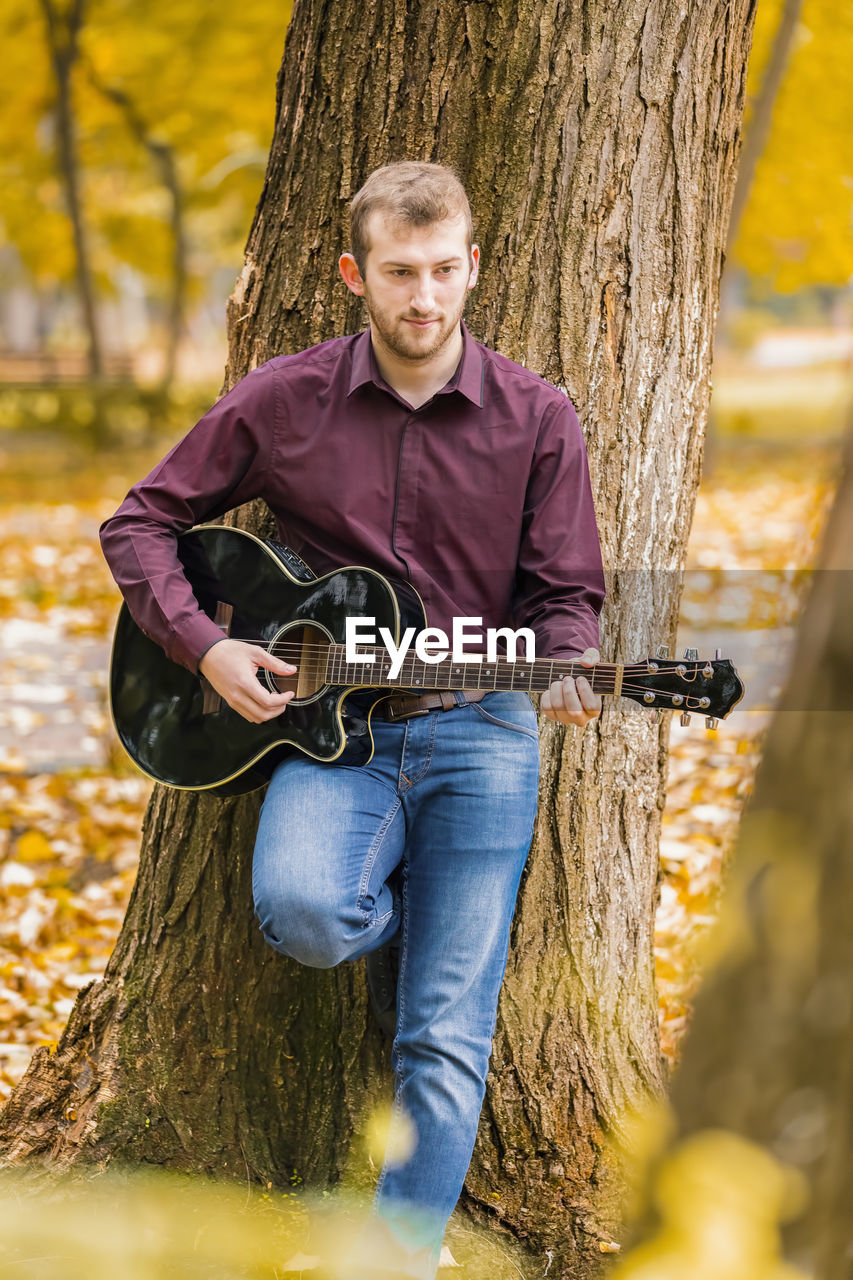 Young man playing guitar while standing by tree in park during autumn