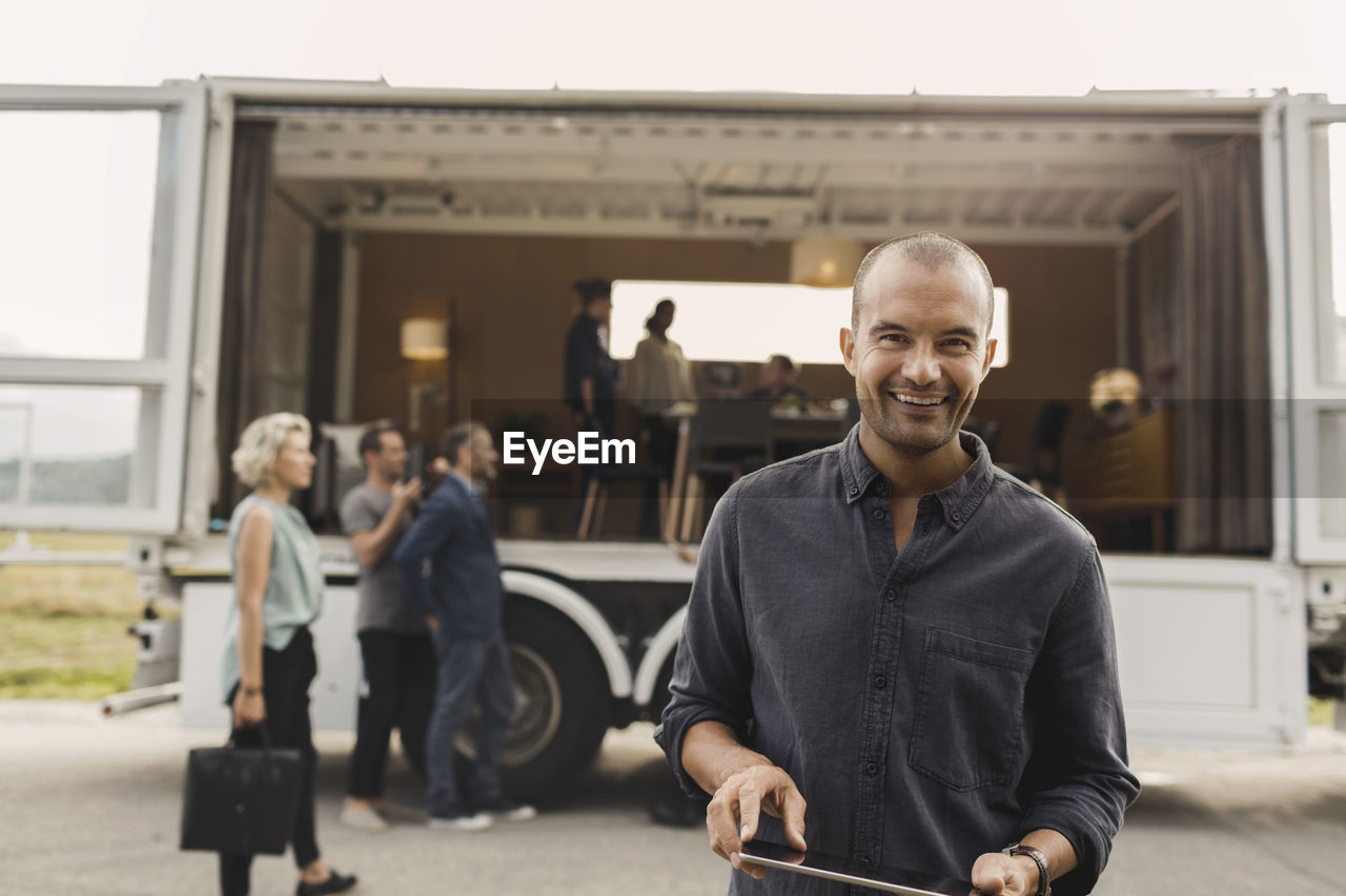 Portrait of smiling businessman holding digital tablet with colleagues and portable office truck on road in background