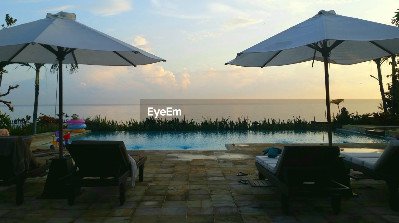 Umbrellas and lounge chairs by swimming pool against sky during sunset