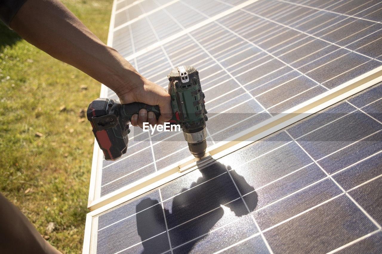 Hand of male engineer using drill on solar panels while working at power station