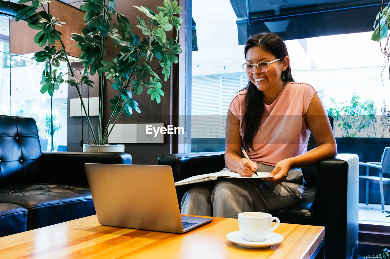 Content young ethnic female student with dark hair in casual clothes and eyeglasses smiling and taking notes in copybook while reading information on laptop and doing homework in cafe