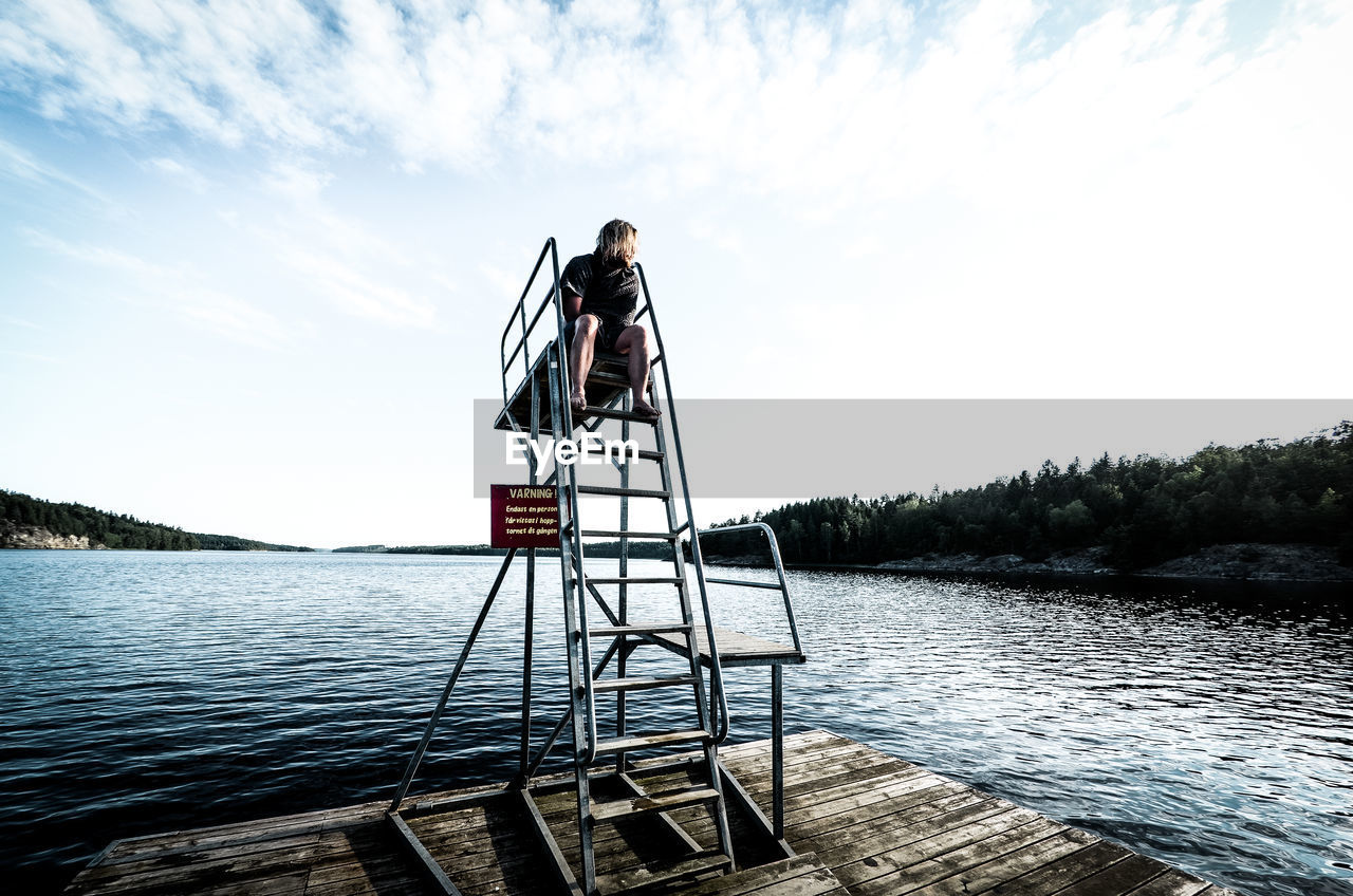 Woman sitting on diving platform by lake against sky