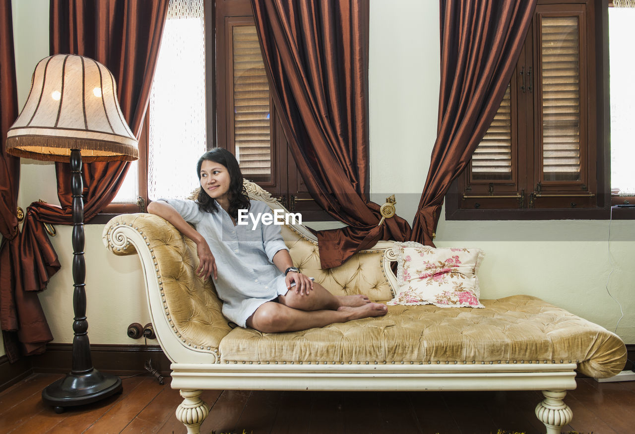 Full length of smiling beautiful woman resting on chaise longue at home