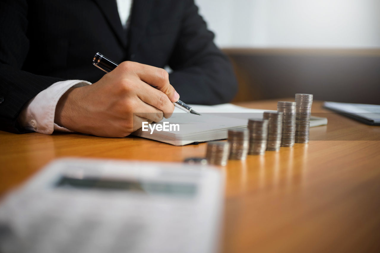 Midsection of lawyer holding pen over book by coins stack on table