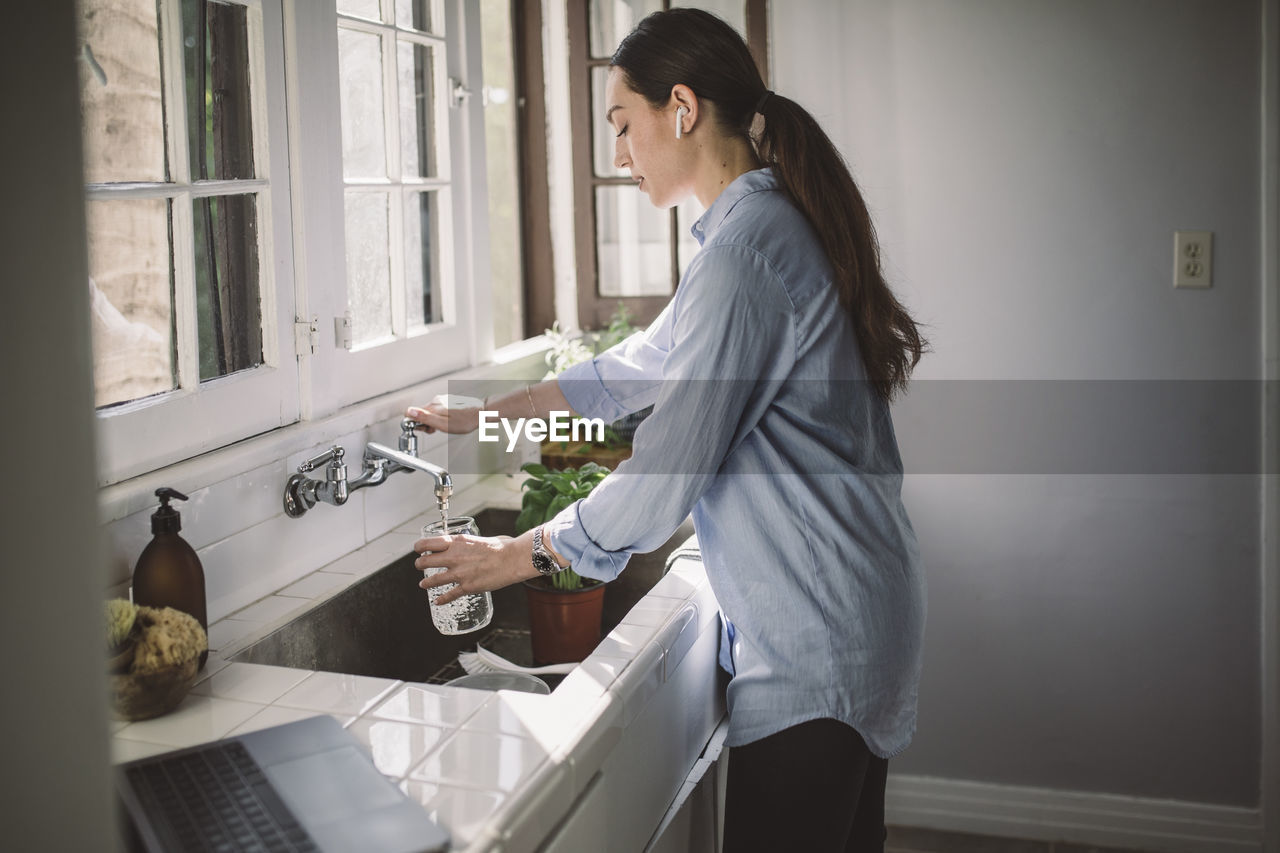 Side view of female business person filling water in jar at kitchen sink