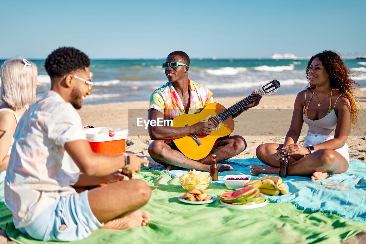 Multiracial men and women in casual clothes drinking beer and eating fruits while sitting cross legged on blanket during picnic on beach on summer weekend day