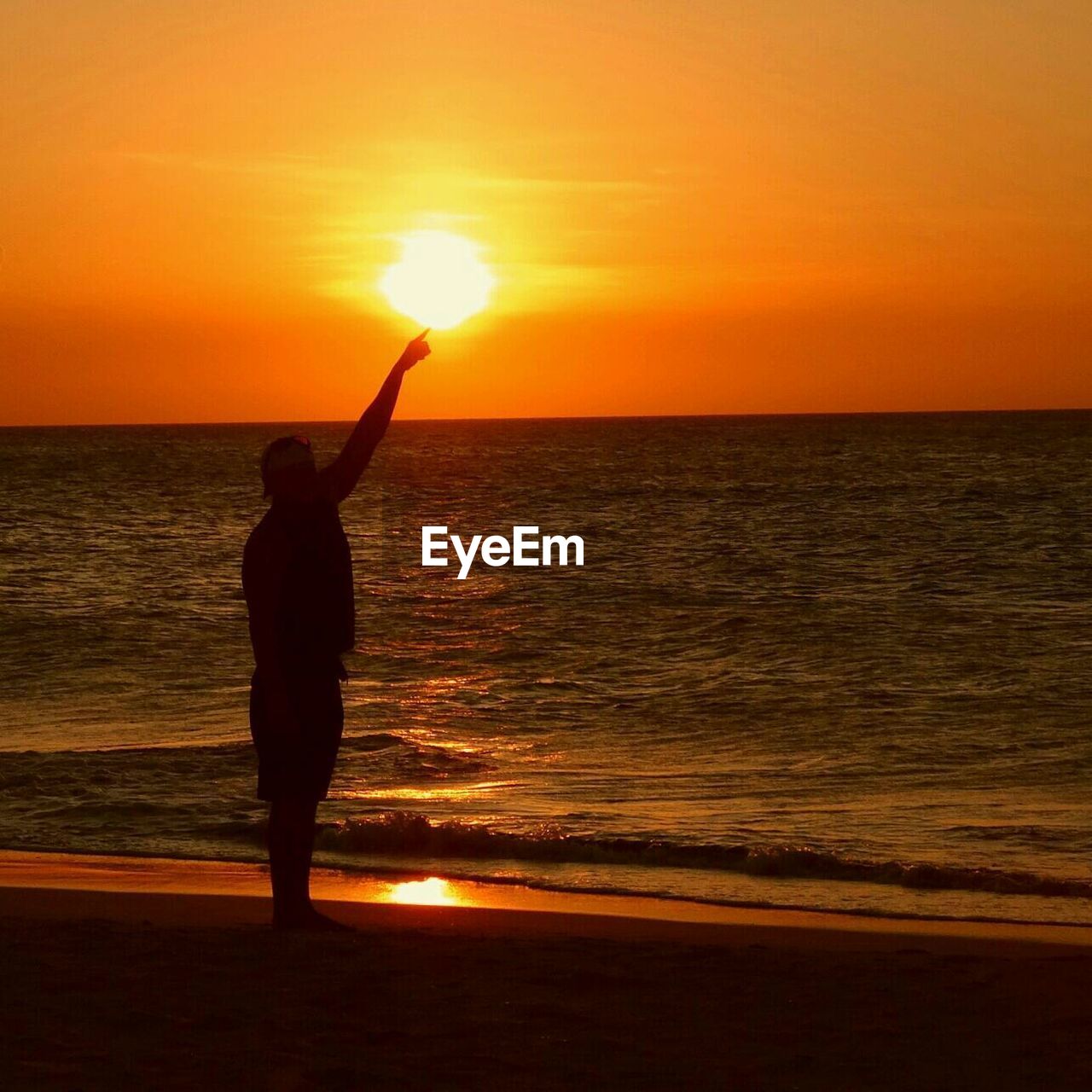Silhouette of man gesturing towards sun at beach during sunset