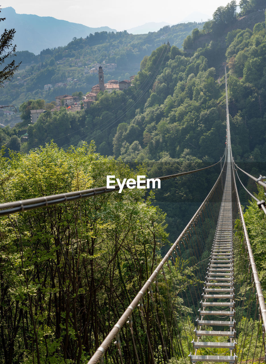 bridge, suspension bridge, plant, mountain, tree, nature, architecture, land, environment, landscape, scenics - nature, built structure, forest, beauty in nature, travel, travel destinations, rope bridge, transportation, sky, no people, tranquility, green, tourism, tranquil scene, outdoors, non-urban scene, day, high angle view, the way forward, lush foliage, foliage, mountain range, rope, railing, water, sunlight, growth