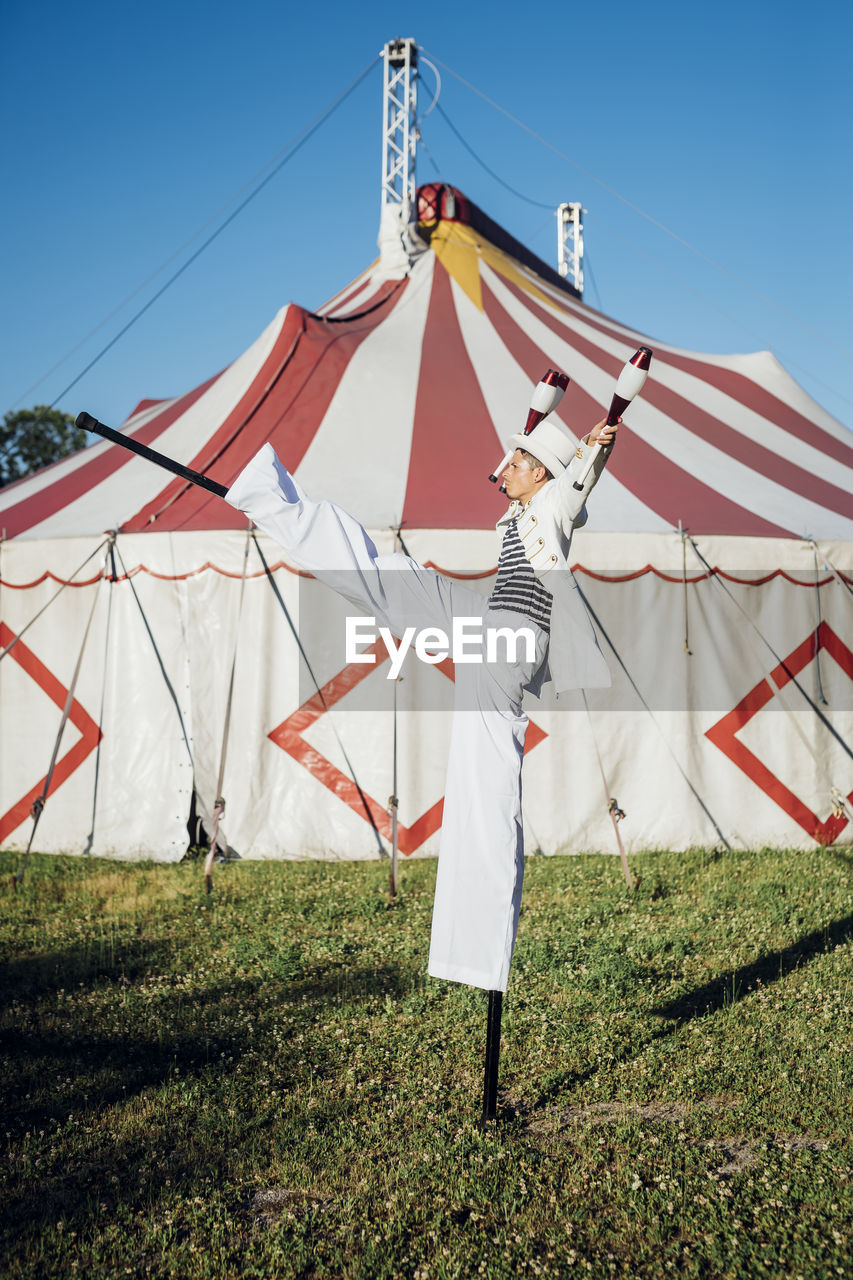 Male artist holding juggling pins while performing with stilts on meadow