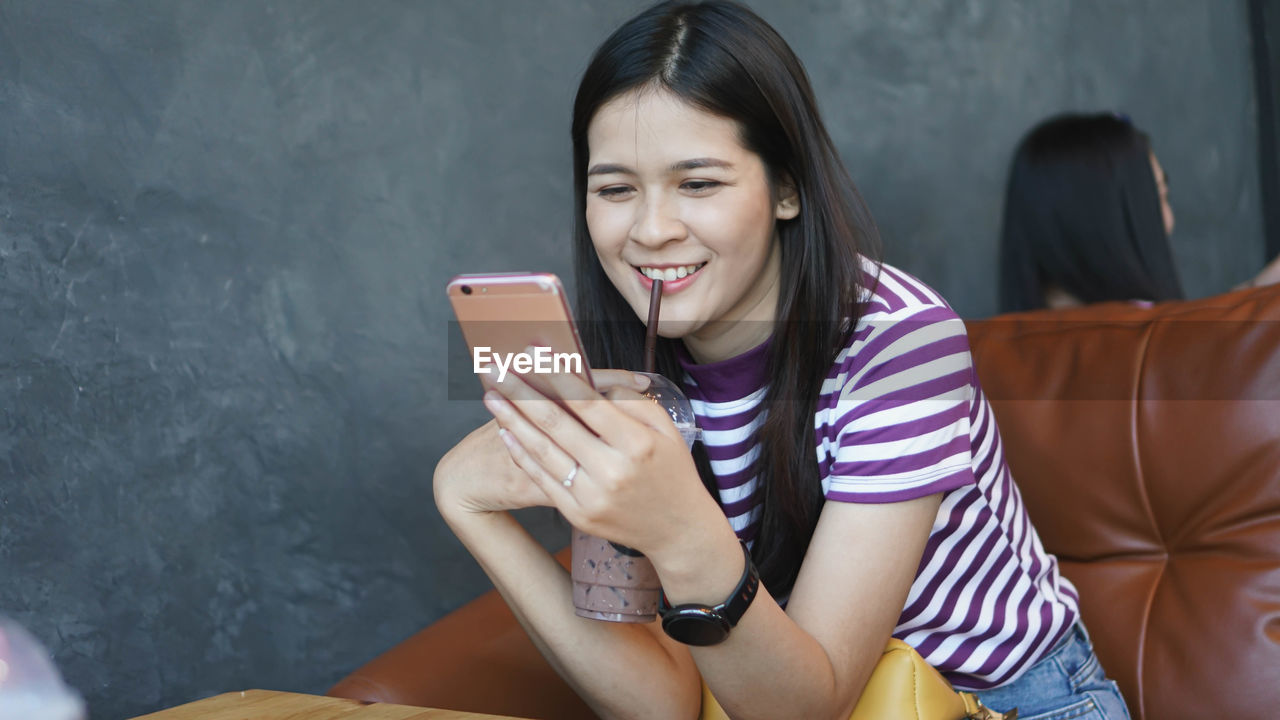 Smiling young woman using smart phone while drinking coffee at table in cafe 