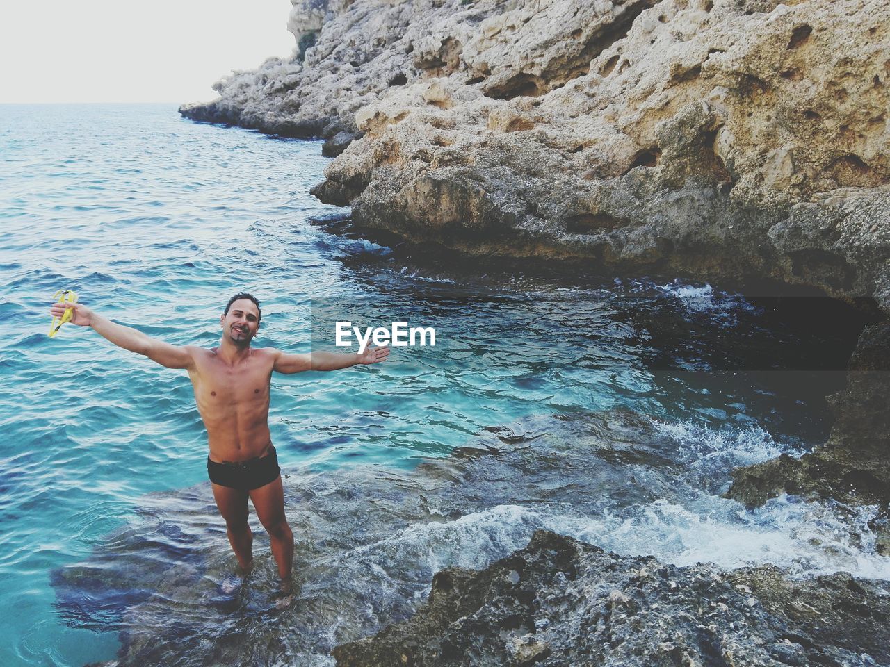 Portrait of young man standing with arms outstretched on rock by sea