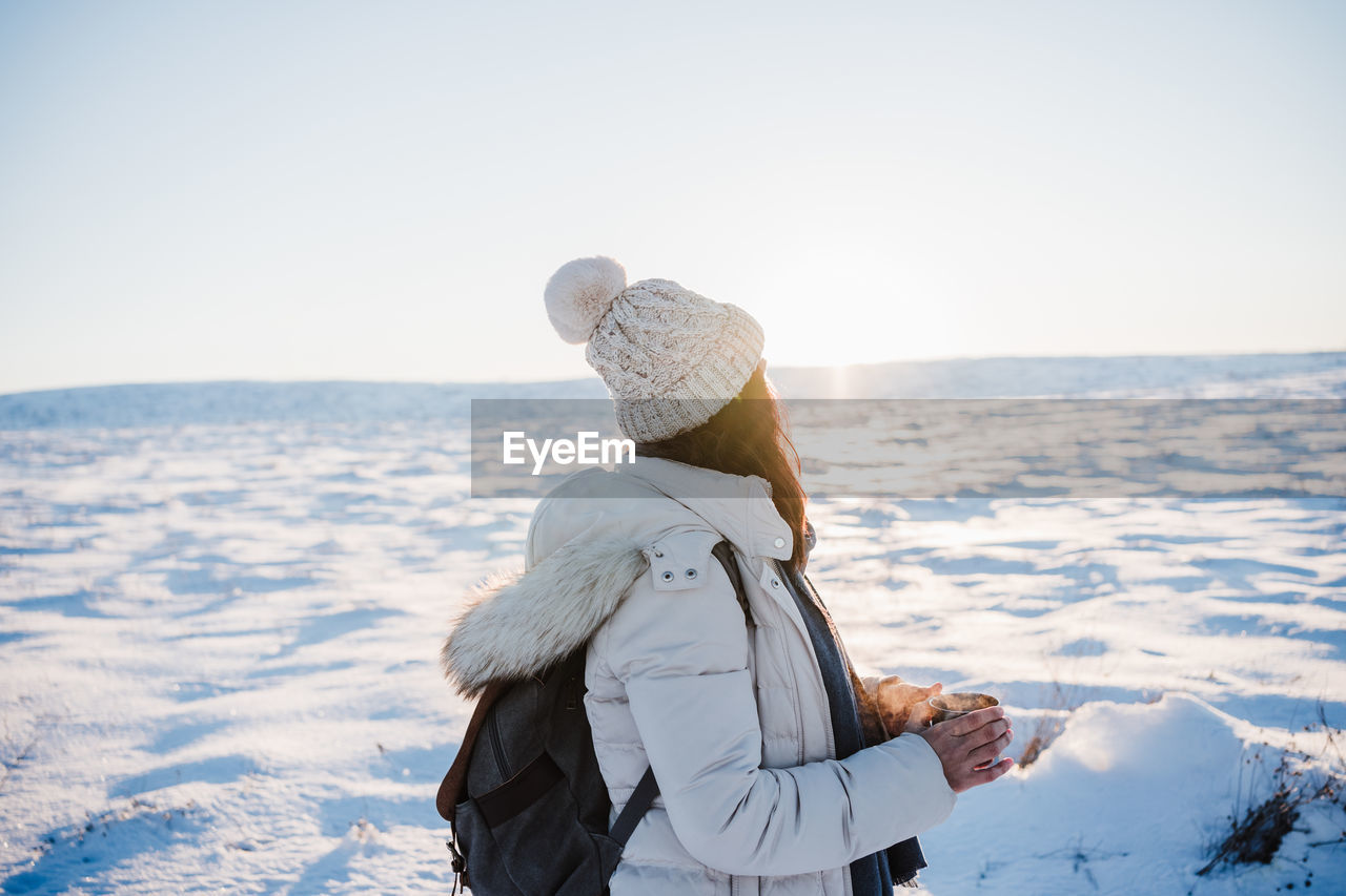Side view of woman looking at snow covered land while holding coffee cup during winter