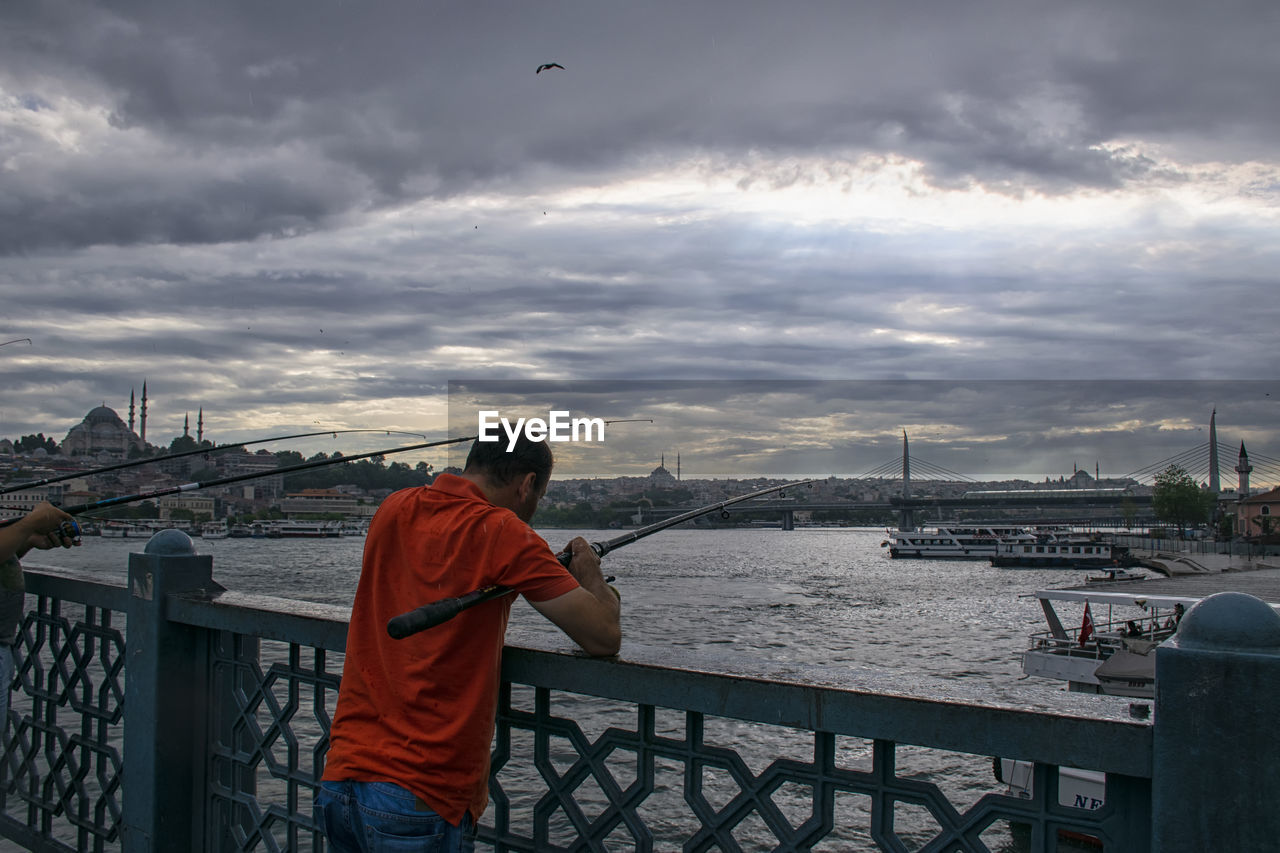 REAR VIEW OF MAN STANDING ON BRIDGE AGAINST SKY