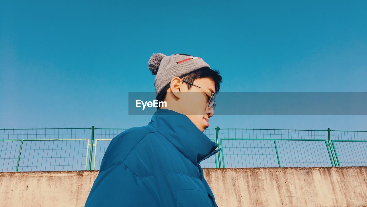 Young man looking down while standing against wall and clear blue sky