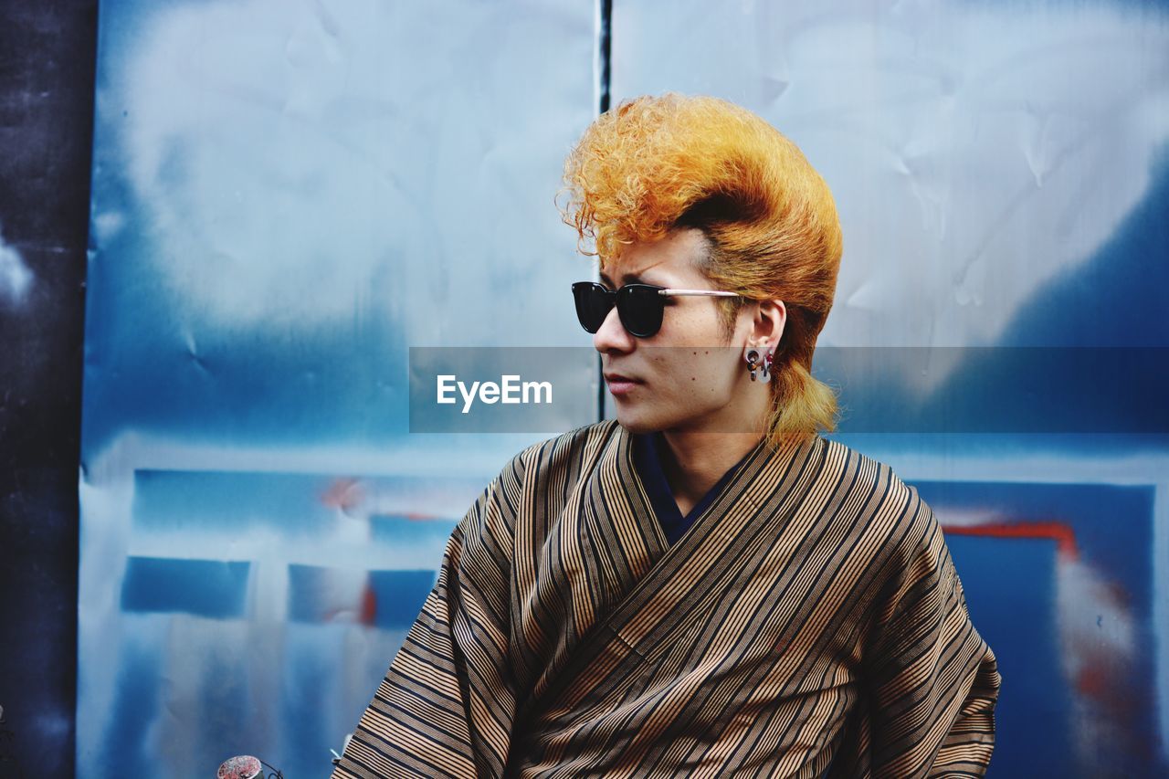 PORTRAIT OF YOUNG MAN WEARING SUNGLASSES STANDING AGAINST BLURRED BACKGROUND