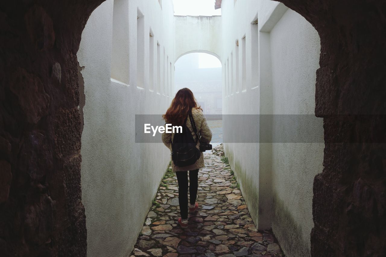 REAR VIEW OF WOMAN STANDING BY WALL IN ALLEY