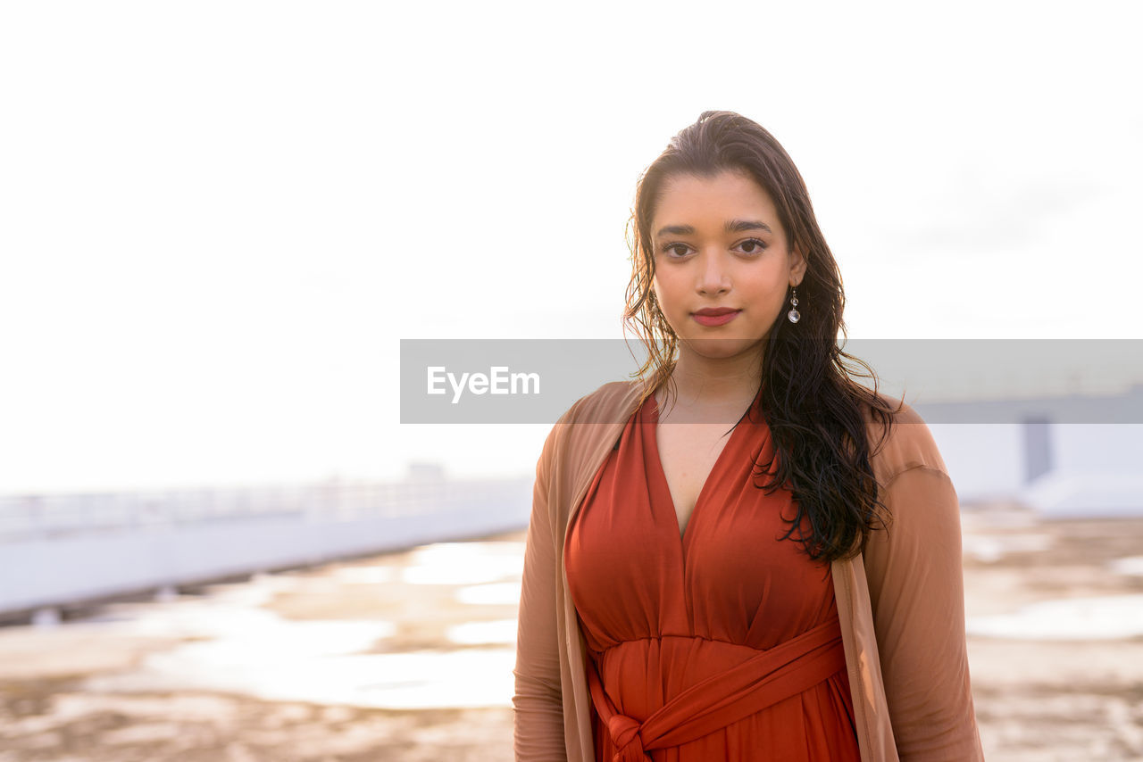 Portrait of young woman standing at beach against sky