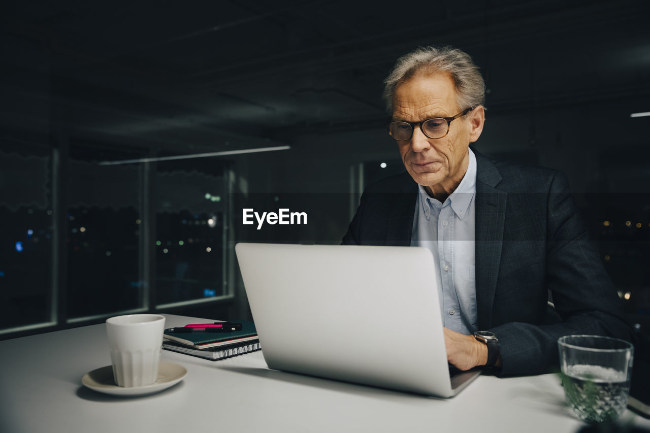 Confident senior businessman using laptop while sitting at illuminated desk in creative office