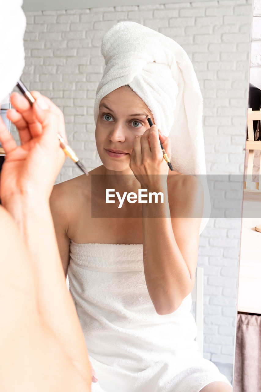 Happy smiling young woman in white bath towels doing morning make up looking at the mirror