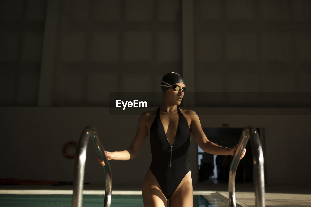 Young beautiful woman leaves the indoor pool by the stairs with black swimsuit, front view