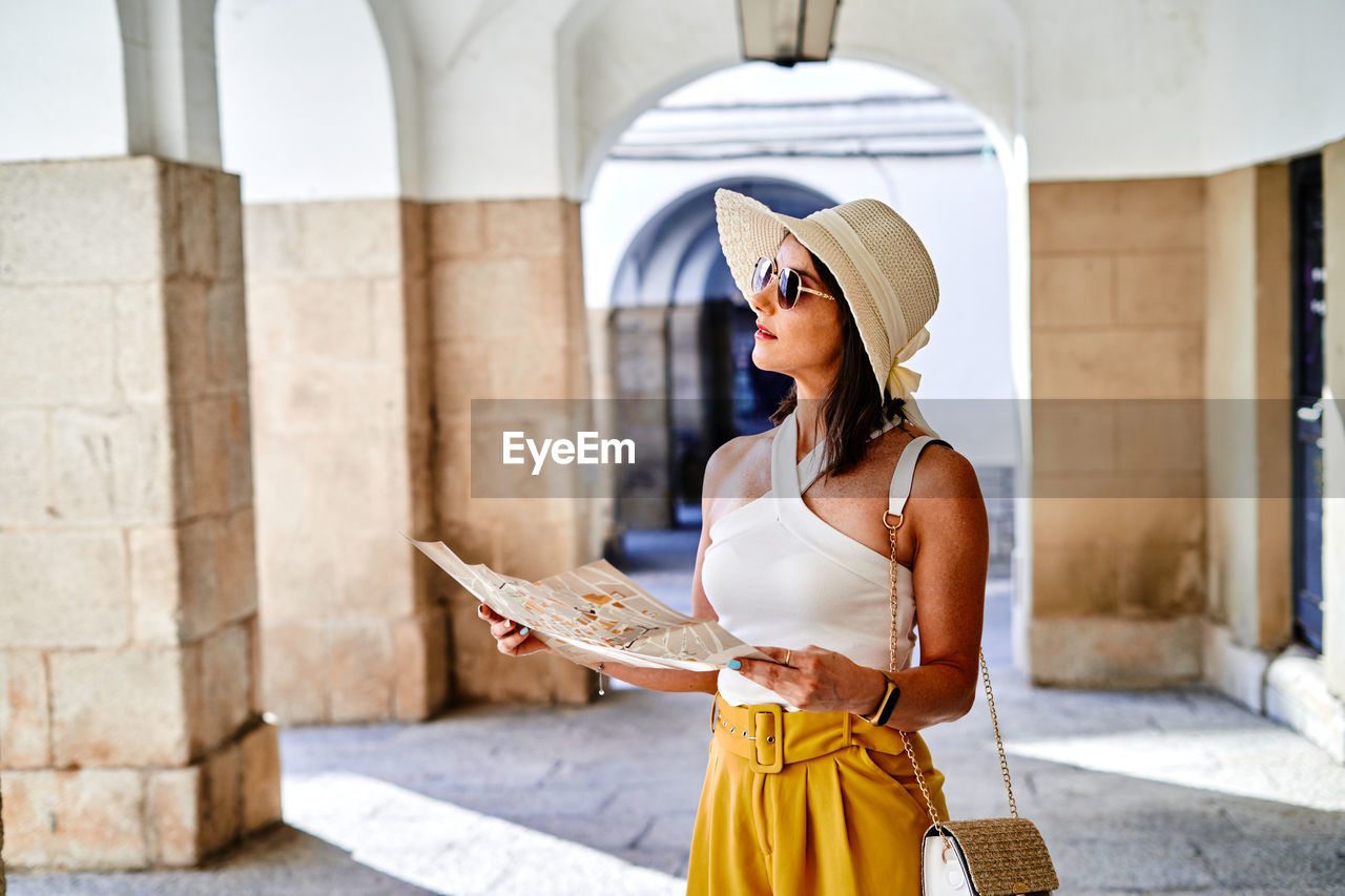 Thoughtful female tourist in hat and sunglasses reading paper map while standing on street during summer vacation and orientating in city