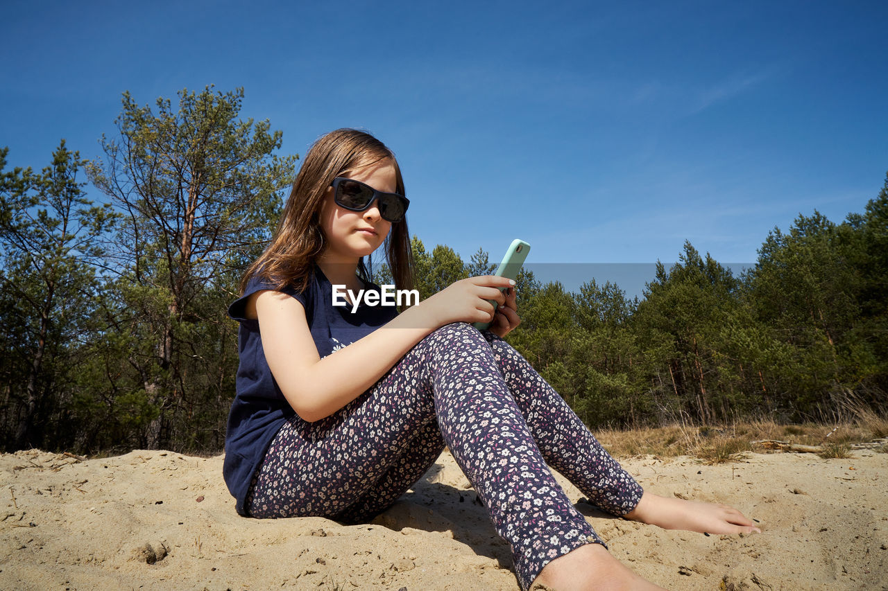 Cute little girl holds phone in hand and sits in summer forest