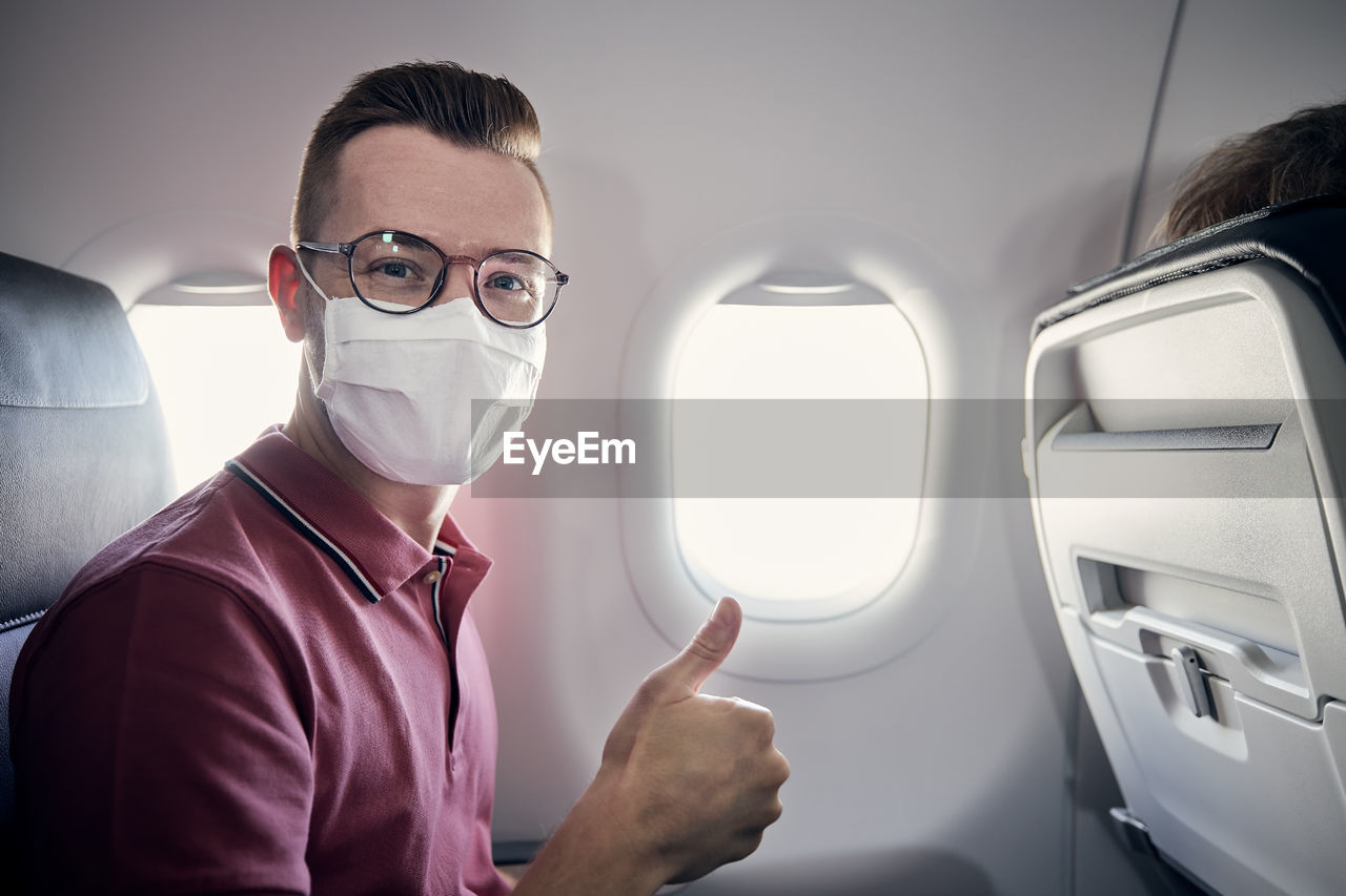 Portrait of happy tourist with face mask during flight. young man showing thumbs up in airplane.