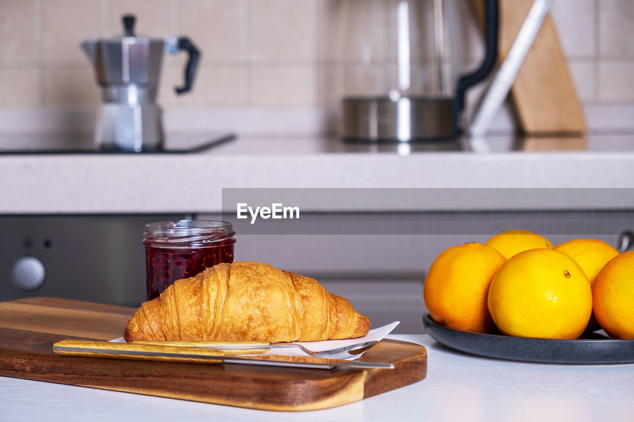 Wooden serving board with croissant and jam and dish of oranges lies on kitchen table in kitchen