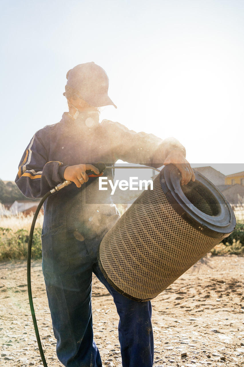 Unrecognizable farmer in workwear and protective respirator mask spraying water from hose to dirty filter of agricultural machine during maintenance work in farm yard