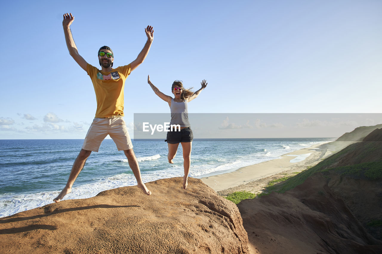 Cheerful couple jumping with arms raised on rock formation at beach