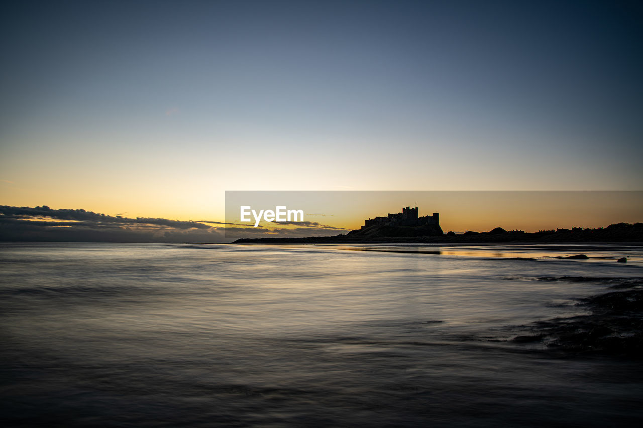 SILHOUETTE BUILDINGS BY SEA AGAINST SKY AT SUNSET