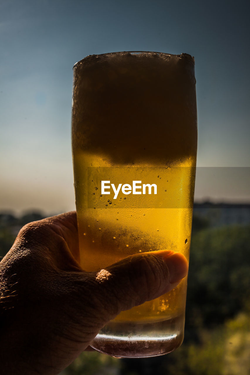 Close-up of hand holding beer glass against sunset