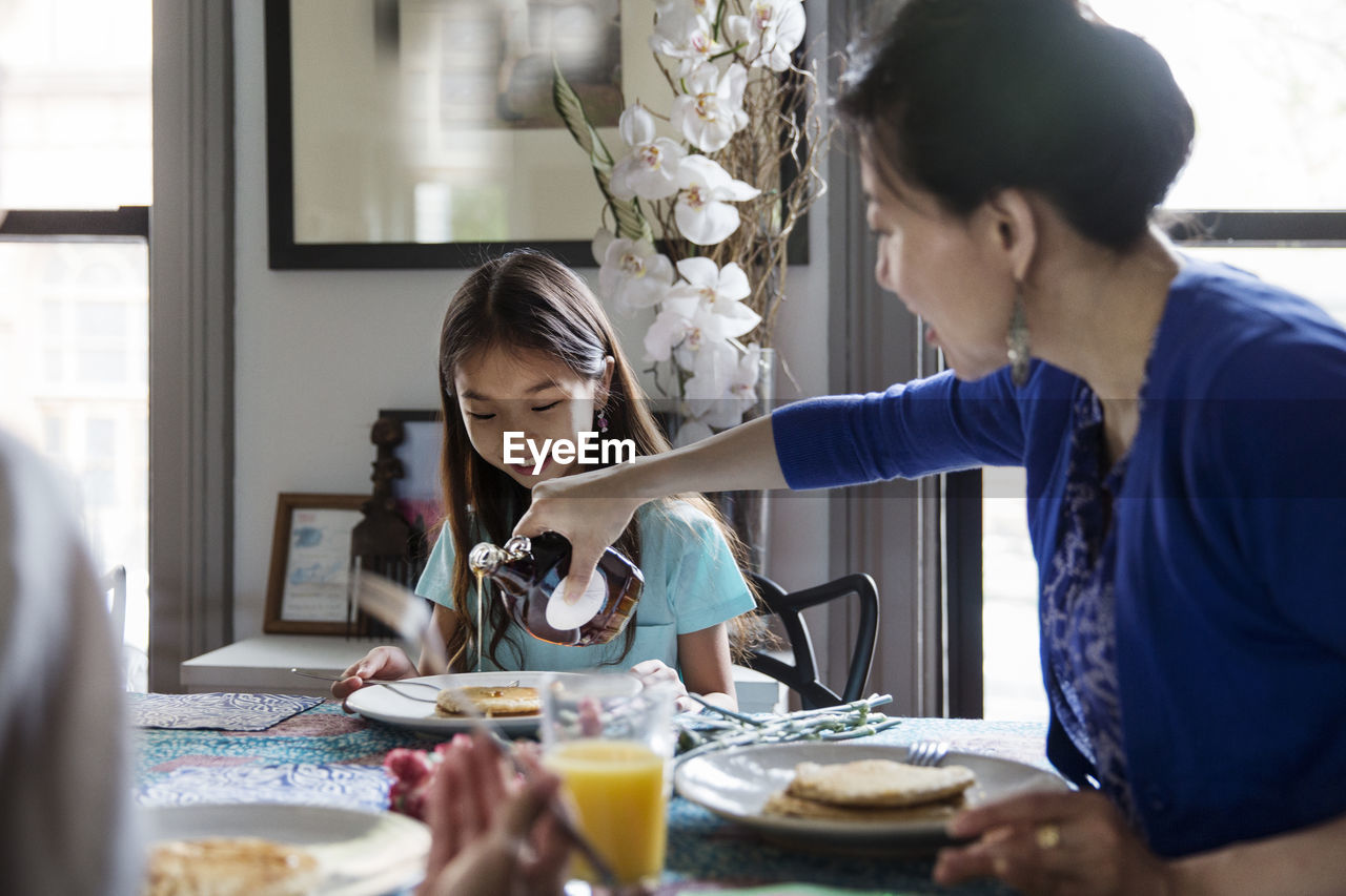 Mother pouring maple syrup on pancakes for daughter during breakfast