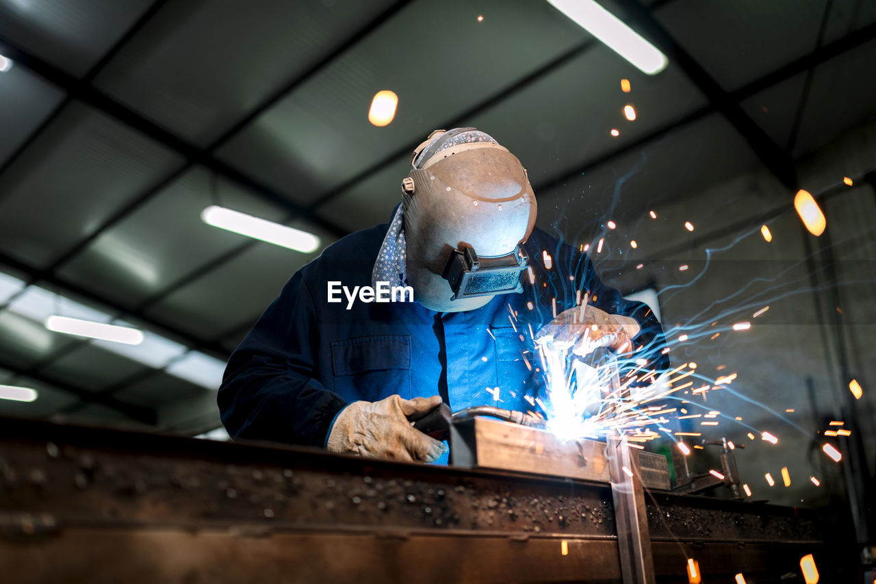 Unrecognizable male employee in protective gloves and helmet using welding machine while working in dark workshop