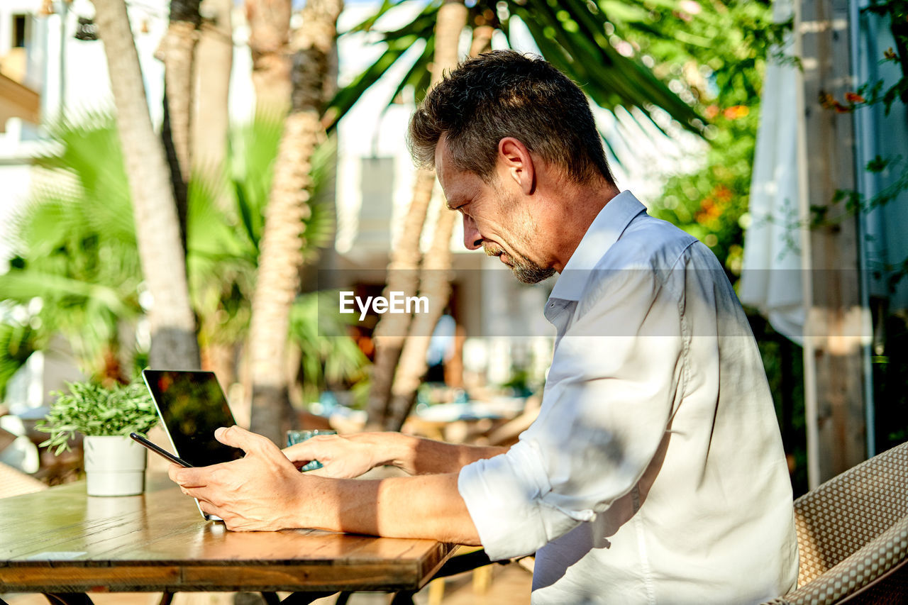 Side view of middle aged businessman in smart casual clothes scrolling data on cellphone while sitting at table with laptop during work in street restaurant