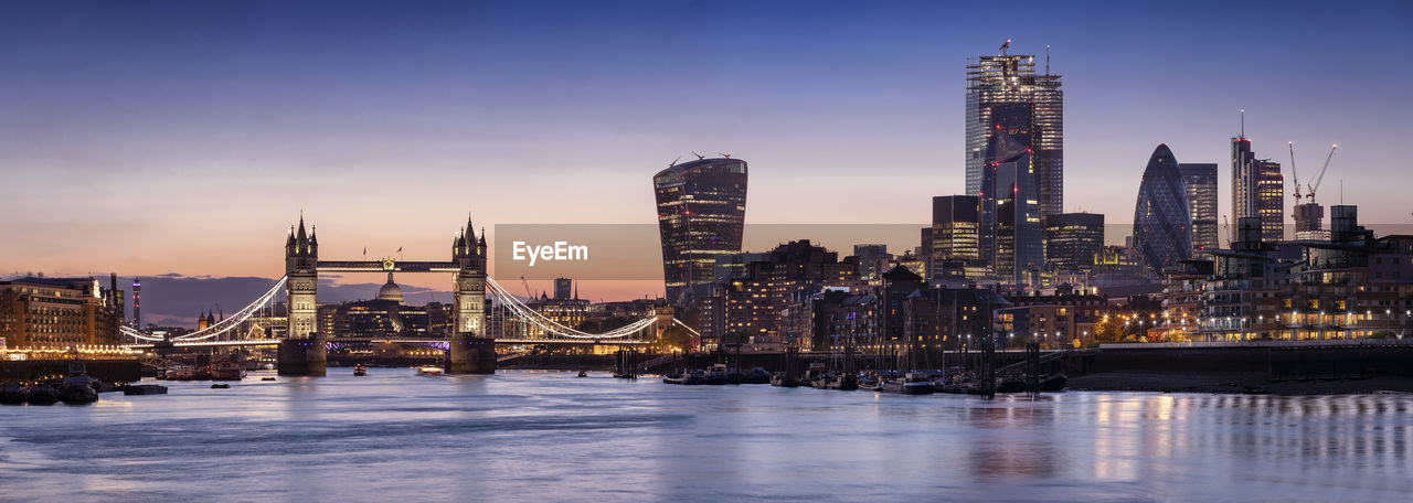 Illuminated city buildings by river against sky