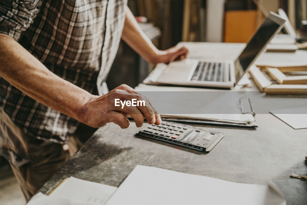 Midsection of male carpenter using calculator on workbench at repair shop