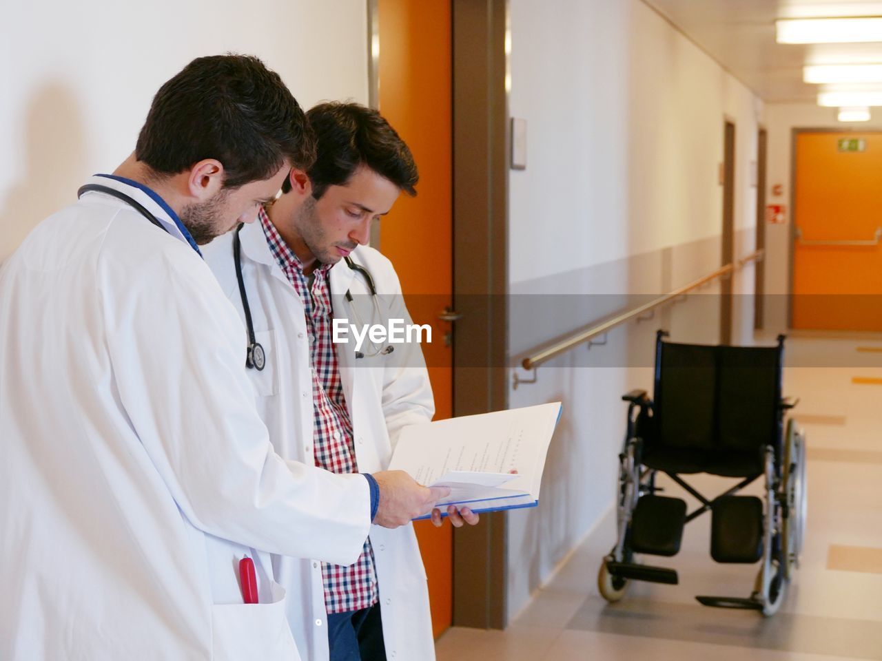 Male doctors discussing over document while standing at corridor in hospital