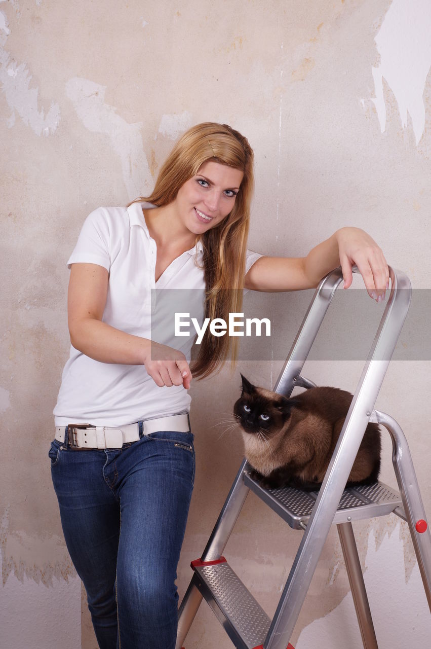 Portrait of smiling beautiful woman with siamese cat on ladder against wall