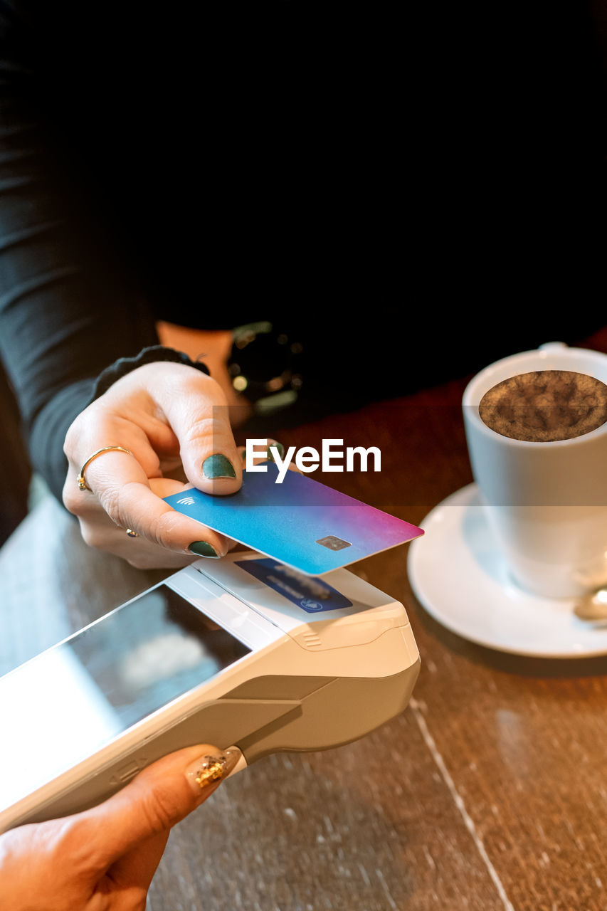 food and drink, adult, one person, coffee cup, cup, coffee, drink, hand, mug, wireless technology, table, indoors, women, holding, lifestyles, cafe, business, refreshment, technology, relaxation, communication, young adult, copy space, leisure activity, crockery, high angle view, female