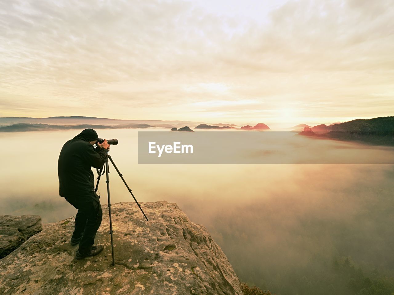 MAN PHOTOGRAPHING ON MOUNTAIN AGAINST SKY AT SUNSET