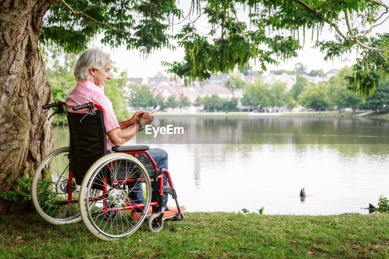 Senior man using smart phone while sitting on wheelchair by lake at park