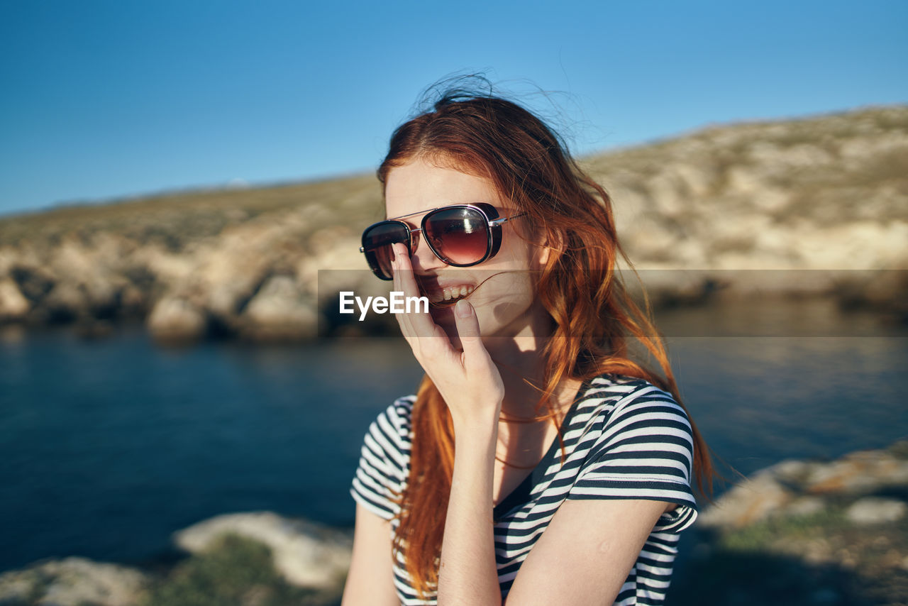 PORTRAIT OF YOUNG WOMAN WEARING SUNGLASSES AT SHORE