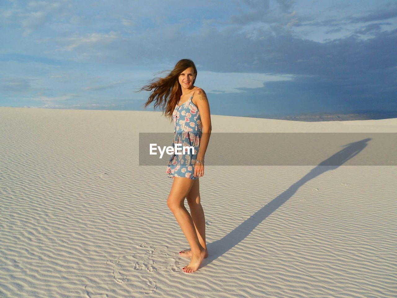 Full length portrait of beautiful woman standing on sand at beach against cloudy sky