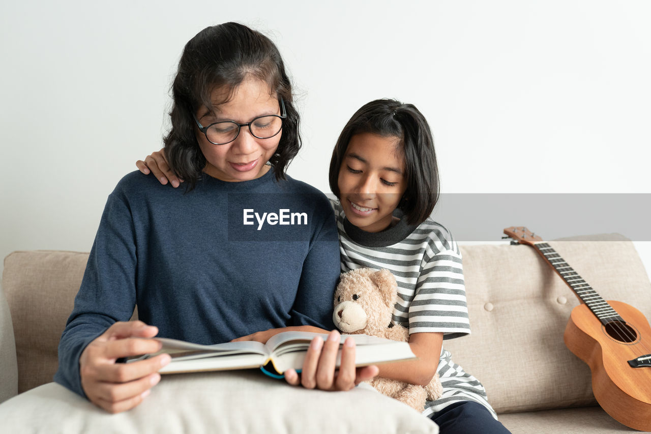 Smiling woman and daughter reading book while sitting on sofa at home