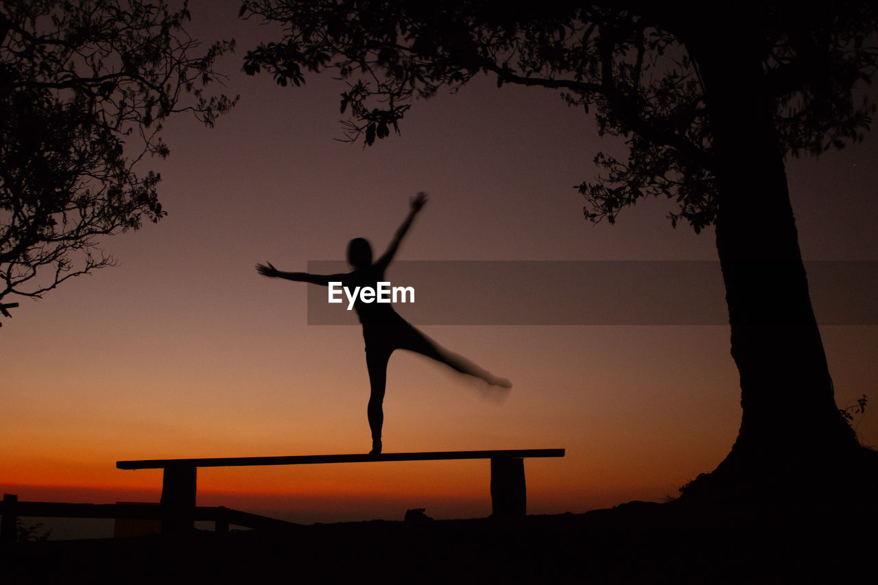 Silhouette woman balancing on bench by trees against sky during sunset