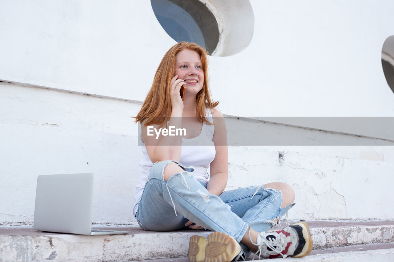 PORTRAIT OF SMILING YOUNG WOMAN USING MOBILE PHONE WHILE SITTING ON CAMERA