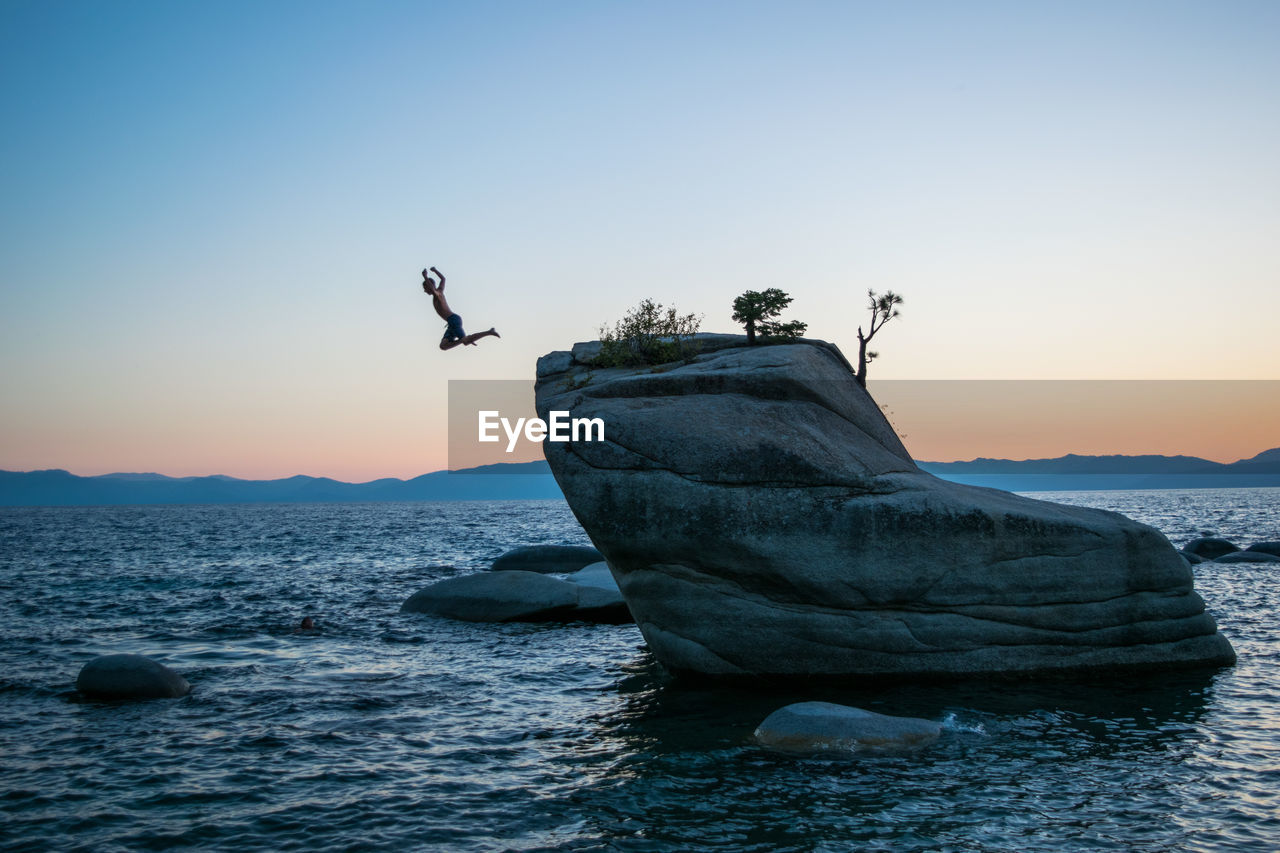 Man jumping off of rock by sea against clear sky