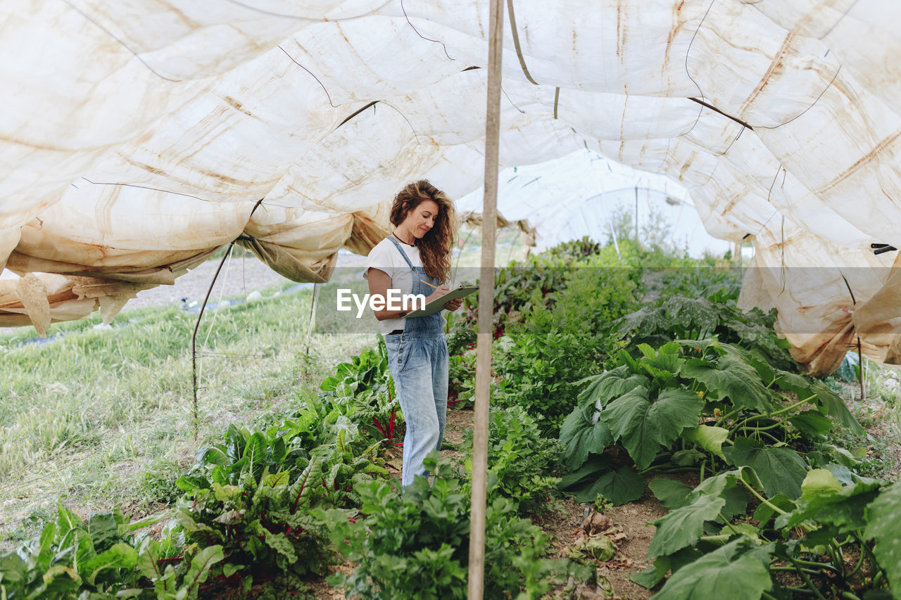 Smiling farm worker with clipboard taking vegetables inventory in greenhouse